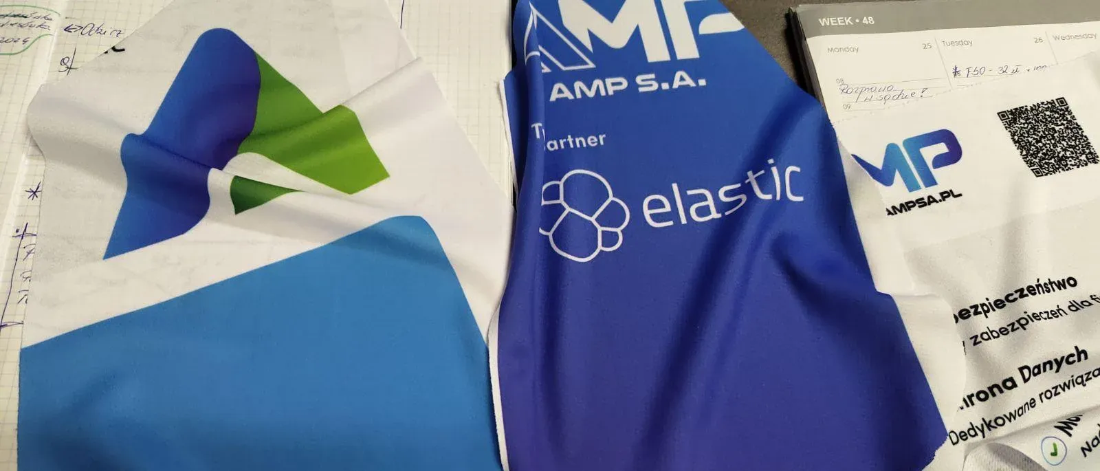 Koszulki firmowe w błękicie i granacie z logo AMP S.A. i logo partnera Elastic, rozłożone na stole.